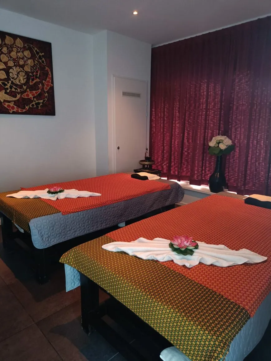 De duokamer van Baipho: twee massagetafels met Thaise kleden, een bordeauxrood gordijn en een handdoek met bloem op elke tafel.